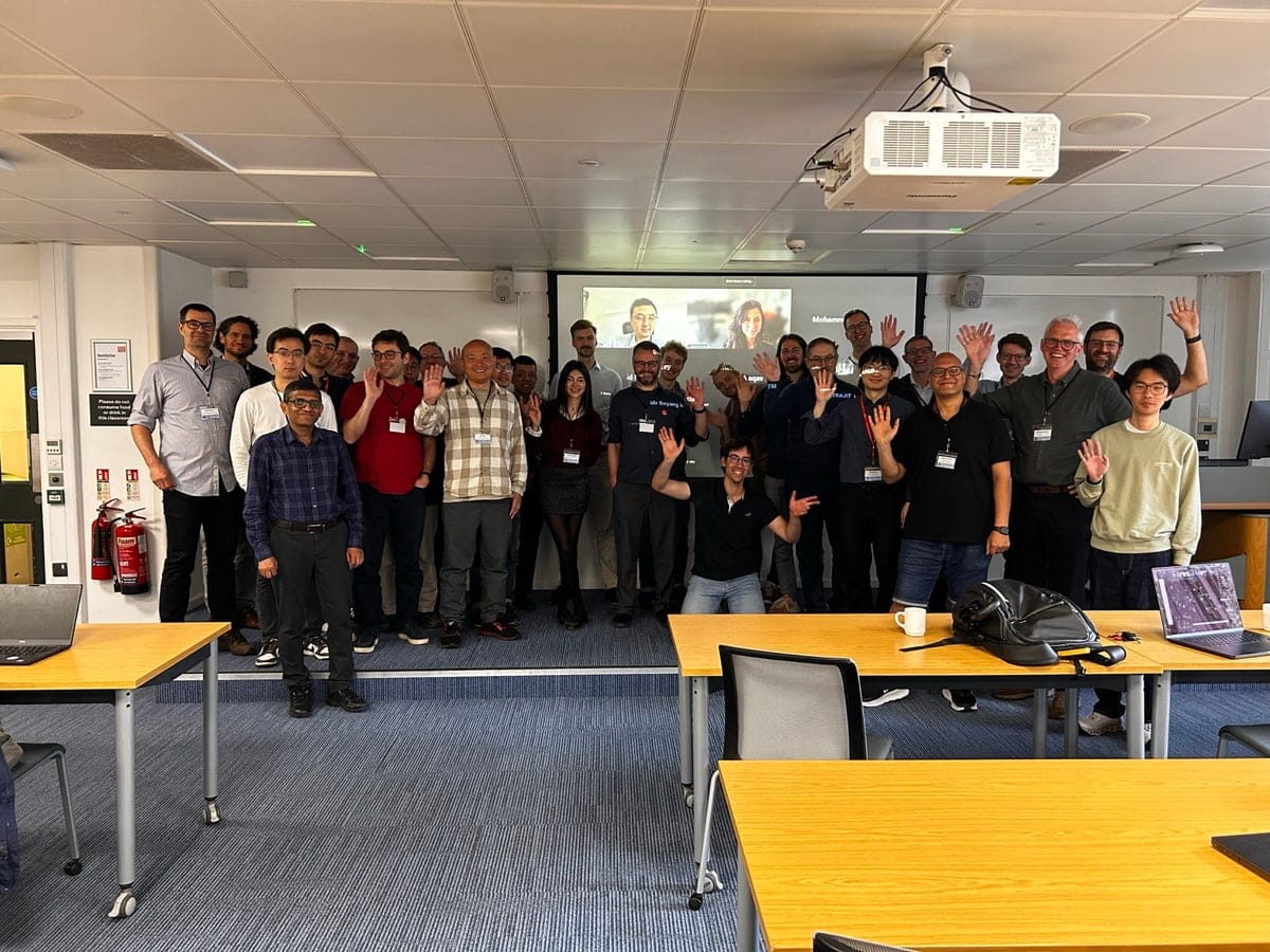 Workshop attendees standing together in a teaching room at King's, waving at the camera, with remote participants visible on the screen behind them.