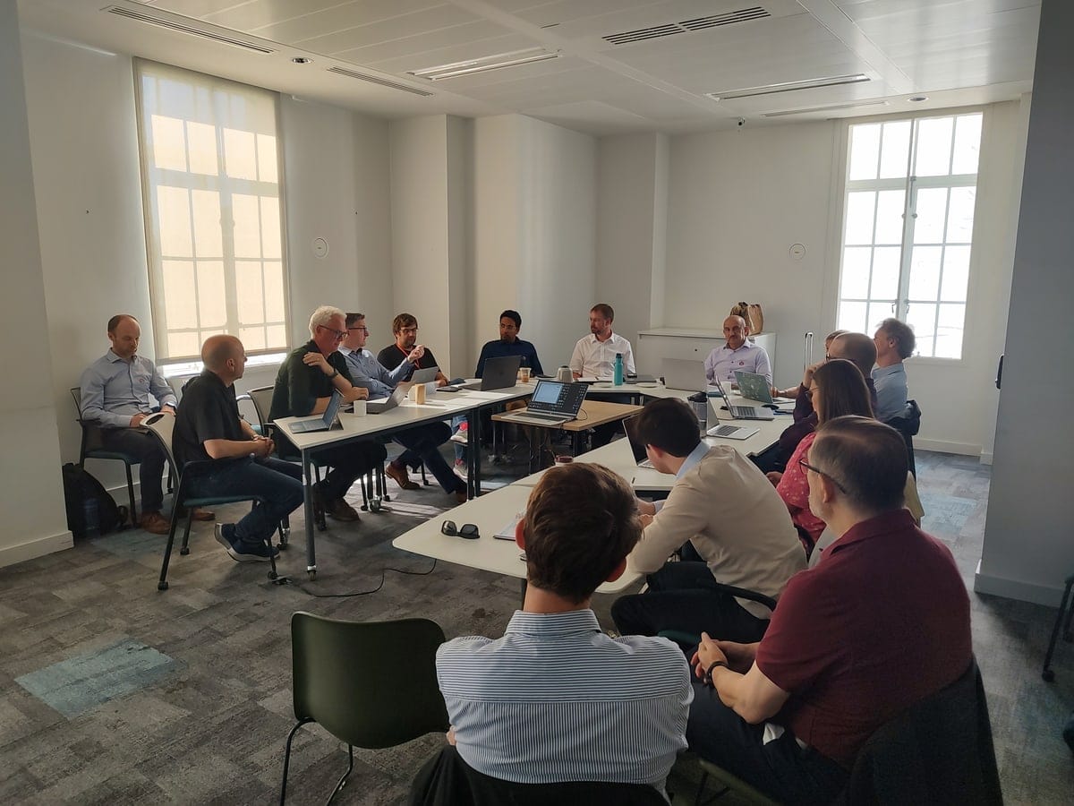 Meeting attendees seated around tables arranged in a square, laptops open, in a room in Bush House.